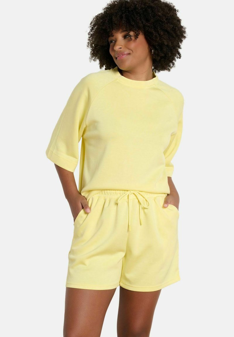 Woman with curly hair wearing a matching yellow short-sleeve top and drawstring shorts, hands in pockets, looking to the side.