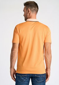 Pomarańczowa koszulka polo z kontrastującym jasnym kołnierzykiem, krótkimi rękawami i gładką fakturą. Noszona z ciemnoniebieskimi dżinsami.