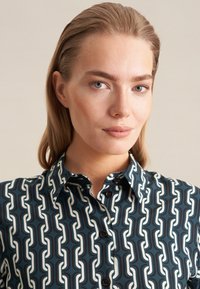 Camicia abbottonata con sfondo di un verde petrolio scuro e un motivo astratto bianco e blu navy ripetuto. Presenta un colletto classico e bottoni scuri.