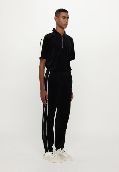Ensemble de survêtement en velours noir avec un col polo zippé, rayures crème sur les épaules et les coutures latérales, assorti à des baskets blanches.