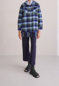 Uld ternet skjortejakke i blå, grøn og mørk marineblå med frynser ved kanten, kombineret med marineblå vidde-bukser og grønne ankelstøvler.