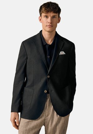 Blazer negro con dos bolsillos frontales y dos botones, llevado sobre una camisa negra con cuello. Un pañuelo de bolsillo estampado realza el blazer.