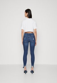 Sötétkék farmer skinny nadrág, elhasználódott szegéllyel. Egy rövid fazonú fehér pólóval párosítva. Hátsó zsebekkel és sima textúrával rendelkezik.