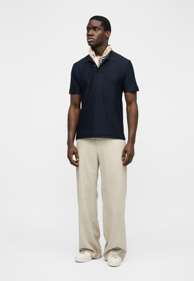 Νέος άνδρας που φοράει μπλε-ναυτικό πόλο με κοντά μανίκια, μπεζ παντελόνι φαρδύ, λευκά αθλητικά παπούτσια και ανοιχτόχρωμο φουλάρι με σχέδια στον λαιμό, στέκεται μπροστά σε απλή φόντο.