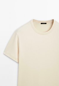 Beige T-shirt met korte mouwen en ronde hals met geribbelde kraag, plat neergelegd met zichtbaar merklabel aan de binnenkant van de halslijn.
