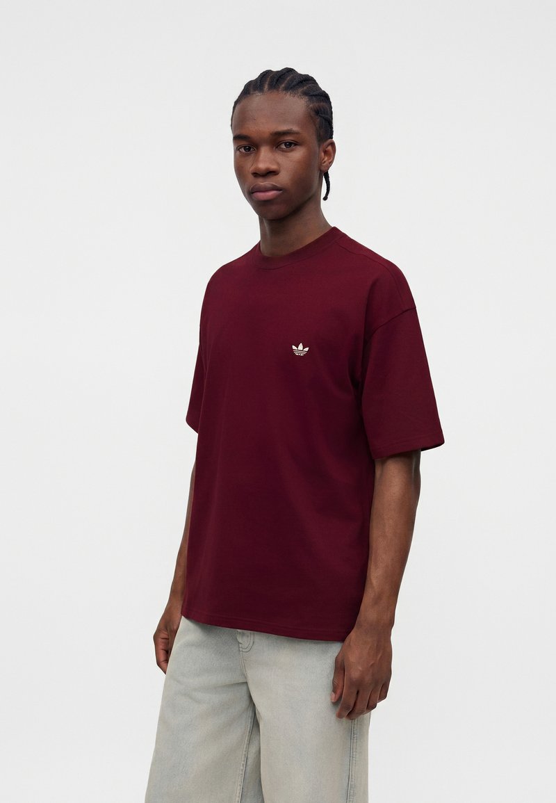 Junger Mann mit geflochtenem Haar trägt ein burgunderrotes Adidas-T-Shirt und hellgraue Hosen und steht vor einem schlichten weißen Hintergrund.
