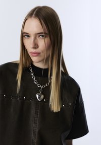 Top in finta pelle nera con maniche corte, caratterizzato da borchie argentate e colletto a costine, abbinato a una collana in argento spessa con pendente a forma di cuore.