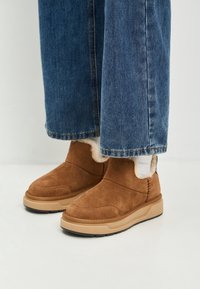 Stivali alla caviglia in suede marrone con suole in gomma beige e rivestimento in pelliccia bianca, indossati con jeans di denim blu e calze bianche su un pavimento bianco.