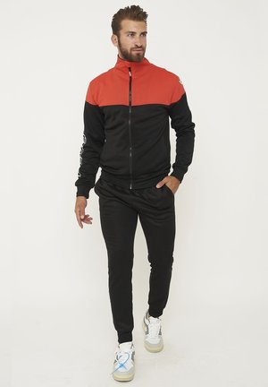Man wearing black and red zip-up track jacket, black joggers, and white sneakers, standing with one hand in pocket, looking right.