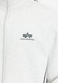 Hellgraue Zip-Jacke aus weichem Stoff. Mit einem schwarzen Logo "ALPHA INDUSTRIES" auf der Brust und dunklen Seitenakzenten.