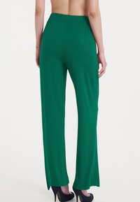 Pantaloni larghi verdi realizzati in un tessuto morbido, dotati di una vita alta e fessure laterali all'orlo. Sono indossati tacchi alti neri.