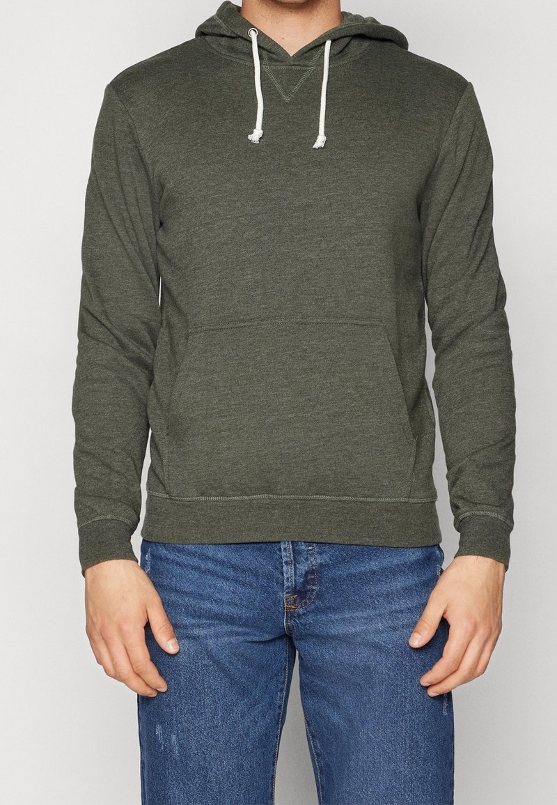 Uomo che indossa una felpa con cappuccio verde oliva con lacci bianchi e tasca frontale, abbinata a jeans in denim blu.