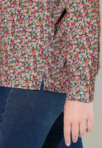 Camicia a fantasia floreale con fiori rosa, rossi e verdi su uno sfondo scuro. Presenta uno spacco laterale e polsini con bottoni. Indossata con jeans.