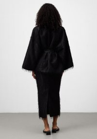 Kobieta z kręconymi włosami, ubrana w czarną, teksturowaną kurtkę z paskiem oraz dopasowaną długą spódnicę, widziana z tyłu, stoi na szarym podłodze.