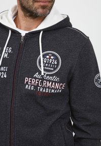 Siva zip-up hoodie s bijelim vezicama, s crvenim i bijelim otiscima teksta. Tekst uključuje "Autentična PERFORMANSA" i okrugle zakrpe na rukavima.