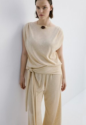 Femme portant un haut beige ample sans manches et un pantalon large assorti noué à la taille, debout contre un mur blanc.