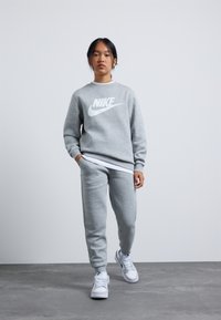 Grijze Nike sweatshirt met wit logo, gecombineerd met bijpassende grijze sweatpants en witte sneakers. Model staat zelfverzekerd op een neutrale achtergrond.