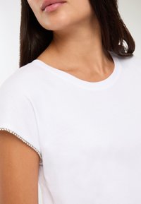 Kobieta w białej koszulce z krótkim rękawem, ozdobionej cekinami na rękawach, z widoczną szyją i częścią twarzy zwróconą w bok.