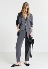 Femme en costume gris à rayures avec gilet et pantalon, tenant un sac à main noir, portant des ballerines noires pointues, regardant vers le bas.