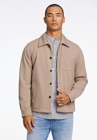 Giacca beige con bottoni, colletto, tasca frontale e bottoni neri, indossata sopra una camicia a righe grigie e bianche, abbinata a jeans azzurri chiari.