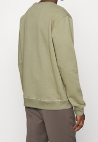 Uomo che indossa un felpa a maniche lunghe verde chiaro e pantaloni marroni, visto da dietro su uno sfondo bianco semplice.