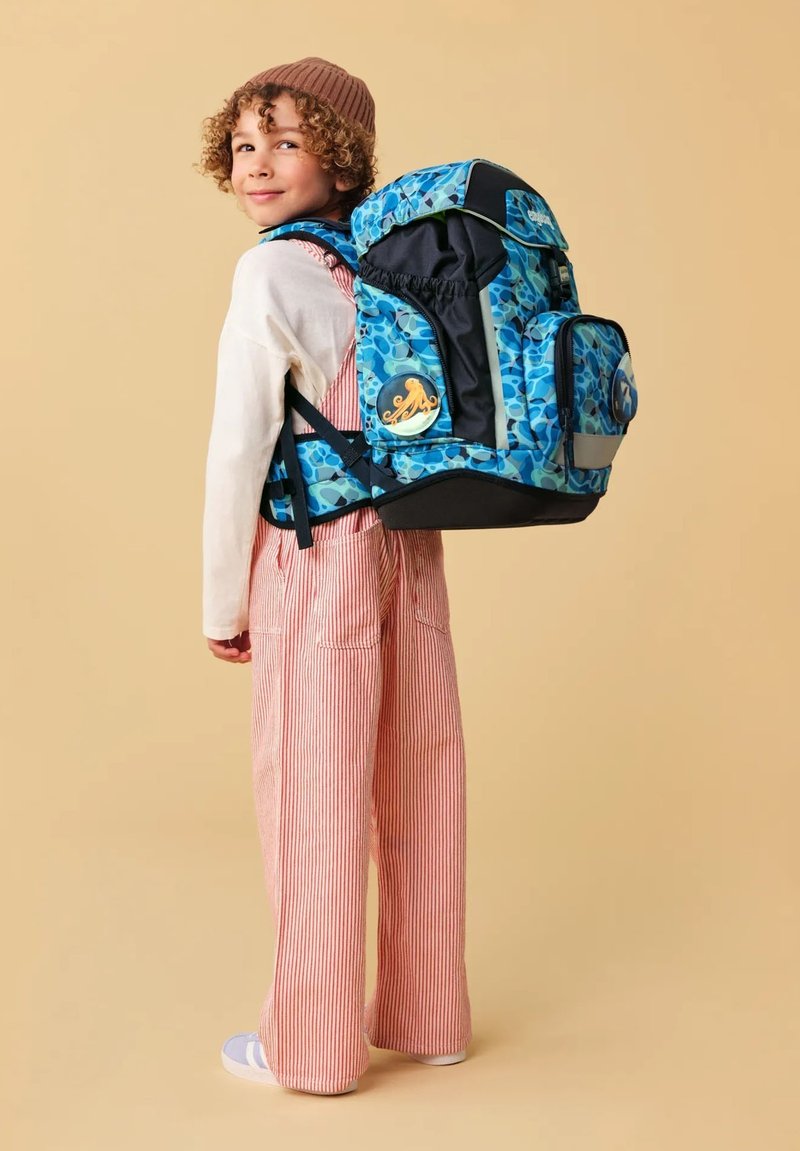Buntes Rucksack mit blauen und schwarzen Mustern, mit einem runden orangefarbenen Oktopus-Abzeichen, getragen von einem Kind in gestreiften Latzhosen.