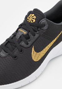Baskets noires et dorées avec lacets noirs, logo doré brodé sur la languette, swoosh Nike doré et semelle blanche sur fond blanc.