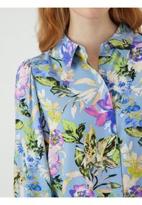 Jasnoniebieska bluzka w kwiaty z kołnierzykiem, zdobiona intensywnymi fioletowymi, różowymi i zielonymi nadrukami roślinnymi; teksturowana tkanina i akcenty w postaci guzików.