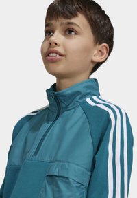 Niño mirando hacia arriba, vistiendo una chaqueta de media cremallera en color teal con detalles en tres franjas blancas en los hombros y las mangas.