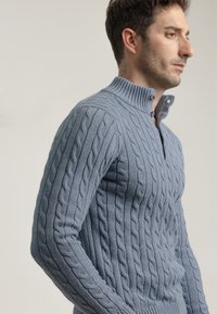 Suéter de punto azul claro con una textura acanalada, que presenta un cuello alto y cierre de dos botones, mostrando un patrón de trenzas a lo largo de las mangas.