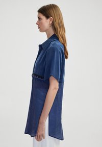Camisa de lino azul de gran tamaño con mangas cortas, cierre frontal de botones y un adorno decorativo de cuentas en la cintura. Textura suave y ligera.