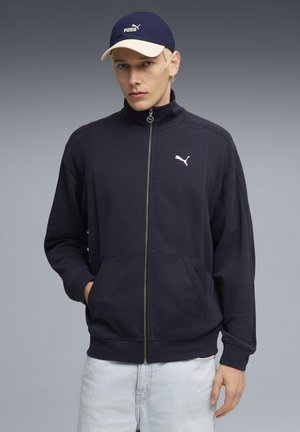Młody mężczyzna ubrany w granatową kurtkę Puma z zamkiem błyskawicznym i kieszeniami kangurzymi, jasne dżinsy oraz granatową czapkę Puma na szarym tle.