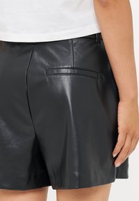 Shorts de cuero negro con una textura suave, que presentan un detalle de bolsillo trasero y un ajuste entallado, mostrados junto a una camiseta corta blanca.