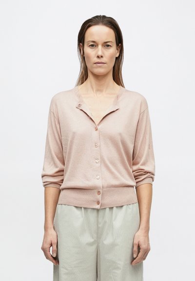 Frau mit glattem braunem Haar, die eine hellrosa geknöpfte Strickjacke und hellgraue, weit geschnittene Hosen trägt, steht vor einem schlichten Hintergrund.