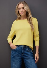 Gelber Strickpullover mit langen Ärmeln und weiter Rundhalsausschnitt, kombiniert mit dunkelblauen Jeans. Glatte Textur, lockere Passform.