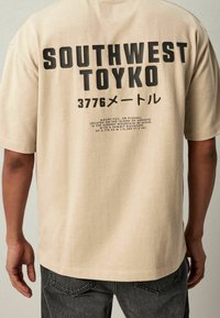 Beige puuvillapaita, jossa on selässä suuri musta graafinen teksti, joka yksityiskohtaisesti kertoo "SOUTHWEST TOYKO" ja Mount Fujista.