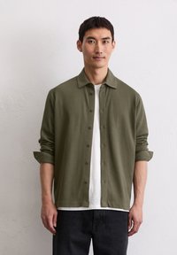 Camicia verde oliva con bottoni, colletto a punta, realizzata in tessuto morbido; presenta maniche arrotolate e delicati dettagli con bottoni. Stile casual.