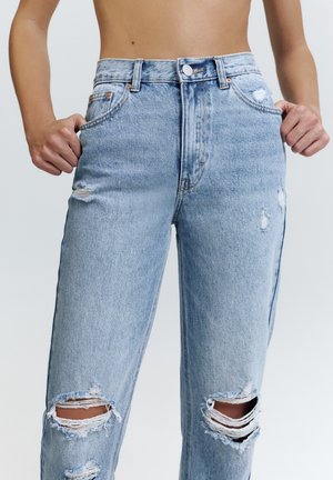 Persoană care poartă blugi rupți, cu talie înaltă, de culoare albastru deschis, cu mâinile în buzunare, evidențiind materialul deteriorat pe coapse și genunchi.