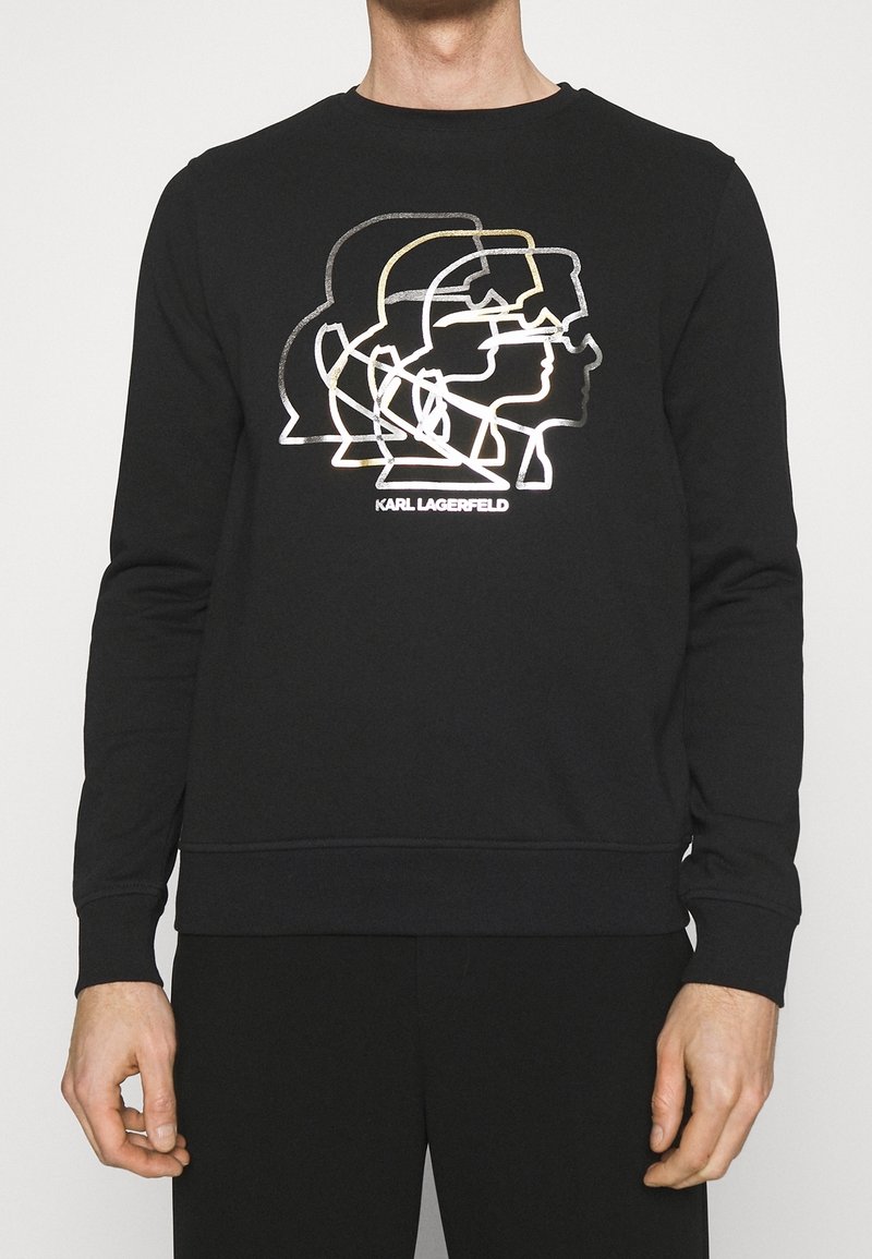 Chemise noire à manches longues avec des profils en ligne blancs et dorés qui se chevauchent et le texte "KARL LAGERFELD" sur la poitrine.
