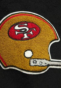 Gesticktes Fußballhelm-Design in Gold und Rot auf schwarzem Stoffhintergrund; mit strukturiertem Stich und kontrastierenden Umrissen.
