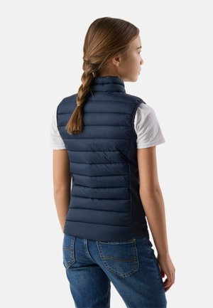 Meisje met gevlochten haar van achteren te zien, draagt een marineblauwe gewatteerde bodywarmer over een wit T-shirt en blauwe jeans tegen een effen lichte achtergrond.