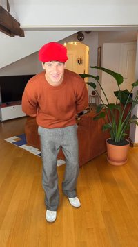 Rode baret, bruine geribbelde trui, grijze geruite broek en witte sneakers. Binnenruimte met een plantenpot en een lichte houten vloer.