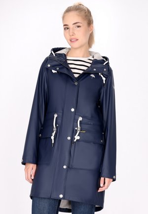 Jonge vrouw draagt een marineblauwe regenjas met capuchon en witte trekkoorden over een gestreept shirt en blauwe spijkerbroek, staand tegen een witte achtergrond.