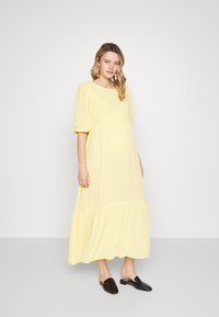 Vestido maxi amarelo com mangas bufantes, corpete franzido e saia em camadas. Tecido suave com textura macia; combinado com sapatos pretos de slip-on.