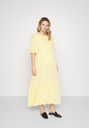 Vestido maxi amarelo com mangas bufantes, corpete franzido e saia em camadas. Tecido suave com textura macia; combinado com sapatos pretos de slip-on.