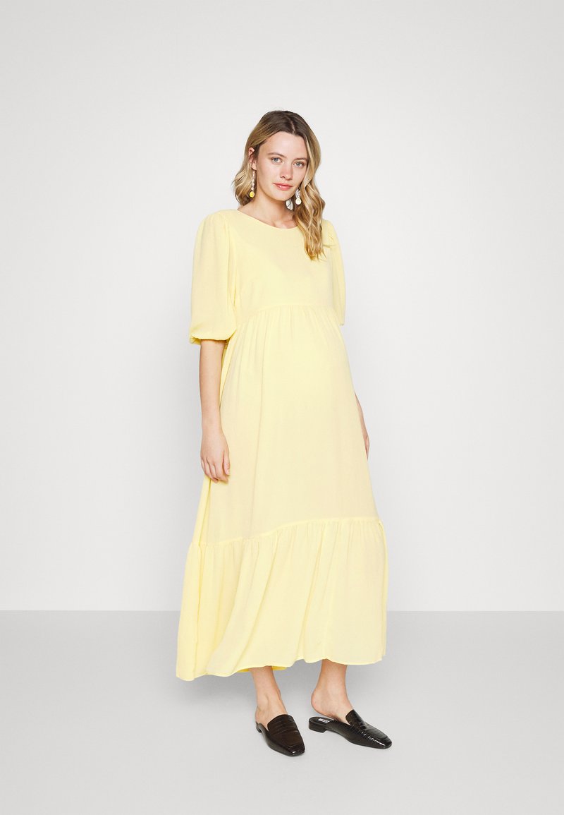 Vestido maxi amarelo com mangas bufantes, corpete franzido e saia em camadas. Tecido suave com textura macia; combinado com sapatos pretos de slip-on.