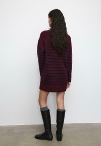 Burgundska haljina džemper s tamnoplavim vertikalnim prugama, pletena tekstura, dugi rukavi i asimetričan rub, uparena s crnim čizmama do koljena.