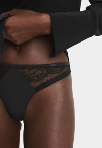 Zwarte slip met een mesh kantpaneel met een bloemenpatroon, een gladde voorkant en een elastische tailleband. Het materiaal is zacht en rekbaar.