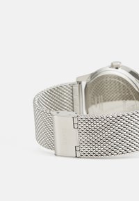 Silbernes Netzarmband mit strukturiertem Gewebe, ausgestattet mit einem Edelstahlverschluss, der mit "GUESS" graviert ist. Runde Uhrenoberfläche auf der gegenüberliegenden Seite sichtbar.