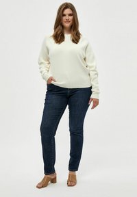 Weißer strukturierter Pullover mit überschnittenen Schultern, kombiniert mit dunkelblauen engen Jeans und hellbraunen Blockabsatzsandalen.